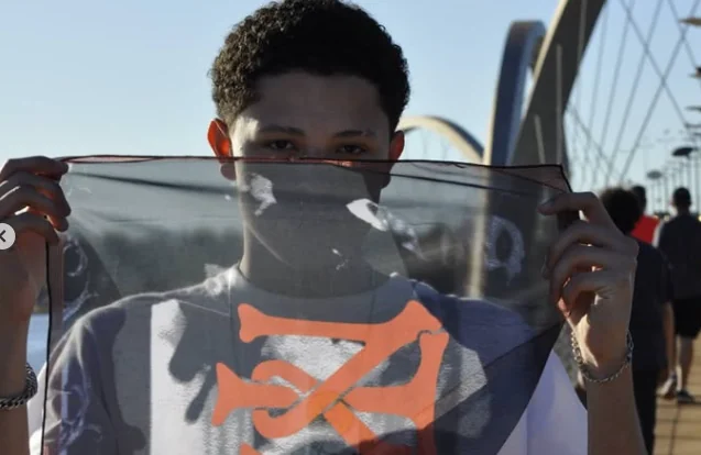 Thalles Leal on a bridge holding a translucent fabric with an orange symbol in front of his face.